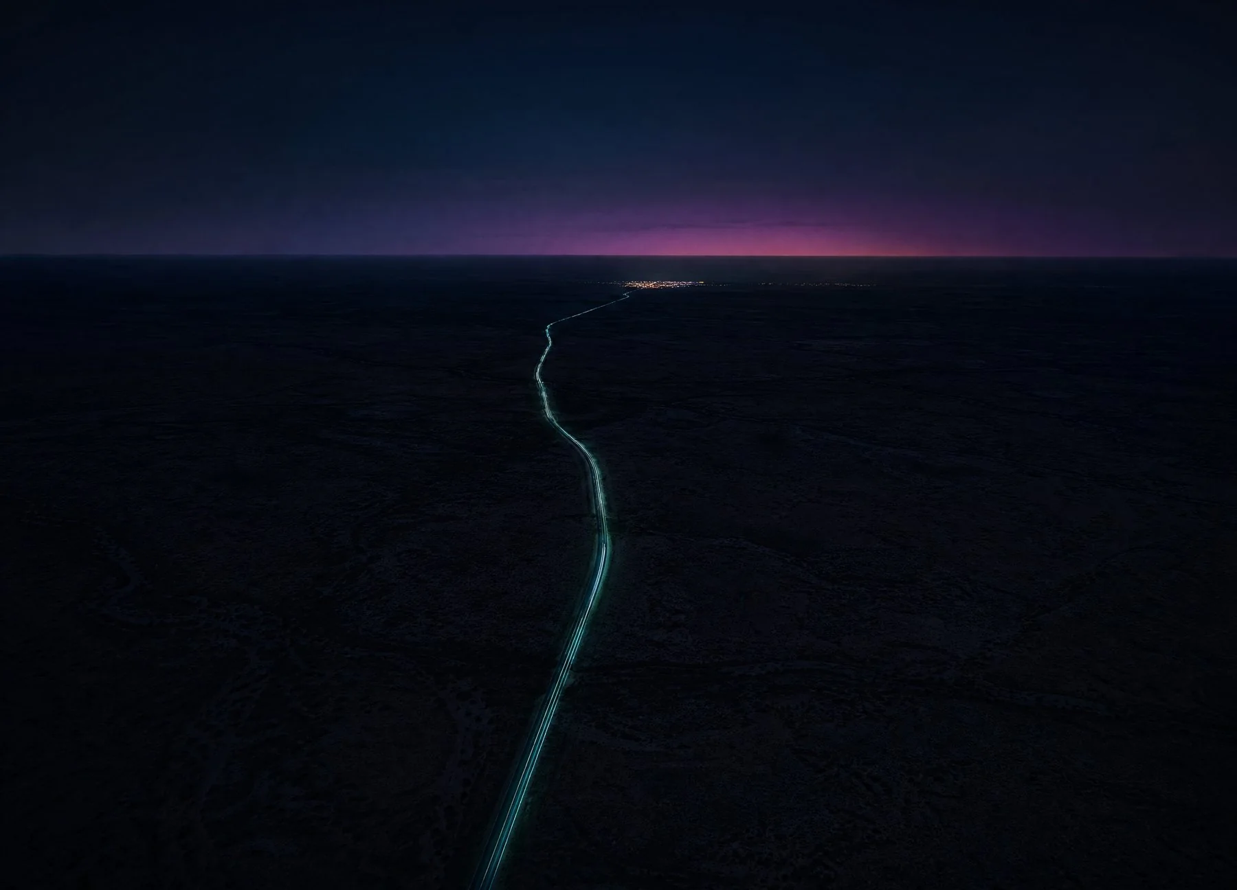 A single road lit in teal running out across dark open country toward a distant town on the horizon