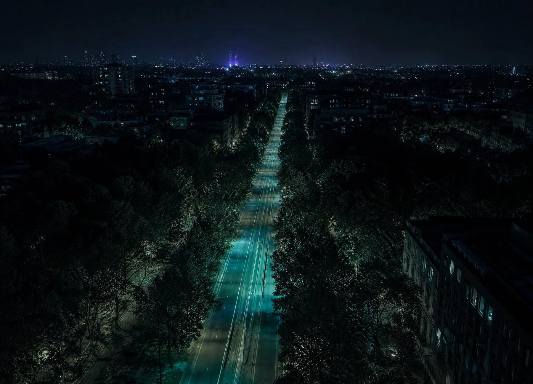 A tree-lined Melbourne boulevard at night from above, its tram tracks lit in teal running away toward the city